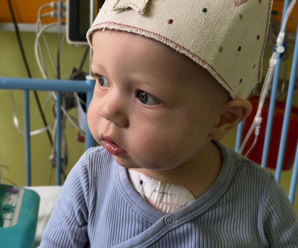 Mały Staś, chłopiec w jasnoniebieskim ubranku i ozdobnym czepeczku, siedzi w szpitalnym łóżeczku, spoglądając w bok z zamyślonym wyrazem twarzy. Przy szyi widoczny jest opatrunek, a w tle elementy aparatury medycznej. O historii Stasia, który wygrał walkę z białaczką, przeczytasz na Poradnik Zdrowie.