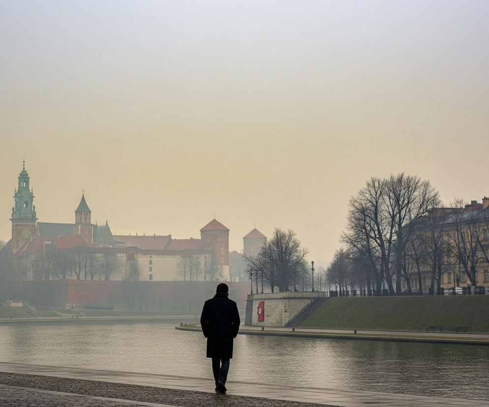 Mężczyzna w ciemnym płaszczu i spodniach idzie tyłem do widza wzdłuż wybrukowanego nabrzeża rzeki, która płynie przez środek kadru. Po lewej stronie rzeki w oddali widoczny jest kompleks budynków z czerwonymi dachami i wysoką wieżą z zielonym, stożkowym dachem, przypominający zamek lub katedrę, częściowo przysłonięty mgłą. Po prawej stronie rzeki znajduje się zielone zbocze z rzędem nagich drzew na szczycie oraz jasnobeżowe budynki mieszkalne w tle. Niebo jest zamglone, o żółtawo-szarym zabarwieniu, dominującym w górnej części obrazu.