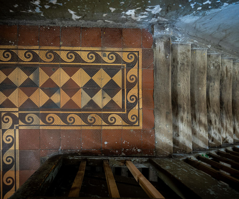 Ozdobna posadzka z płytek ceramicznych obok drewnianych schodów w kamienicy. O wystawie na Pradze przeczytasz w Architektura.