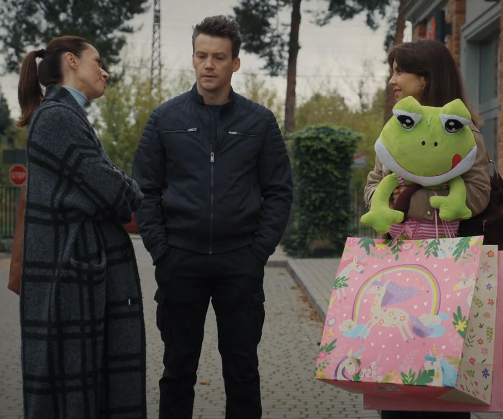 Trzy osoby rozmawiają na ulicy. Kobieta z lewej, Martyna, w szarym płaszczu w kratę, uśmiecha się do mężczyzny w czarnej kurtce, Jakuba, stojącego pośrodku. Kobieta z prawej, Aneta, trzyma pluszową żabę i kolorowe torby z zakupami. Scena z serialu M jak miłość. Więcej o losach bohaterów przeczytasz na Super Seriale.