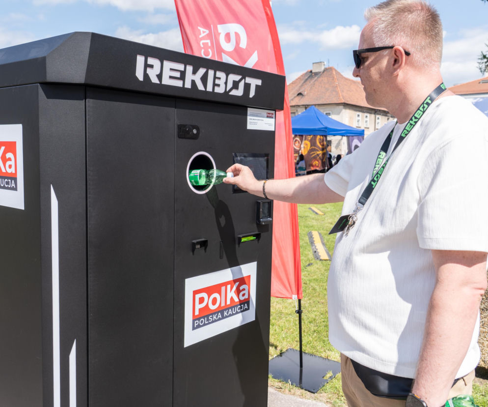 Mężczyzna w białej koszulce i okularach przeciwsłonecznych wkłada zieloną butelkę do Zwrotomatu firmy Rekibot z logo PolKa Polska Kaucja na tle zielonego trawnika i nieba. Nowe Zwrotomaty ułatwią oddawanie butelek w Polsce. Więcej o innowacjach na Super Biznes.