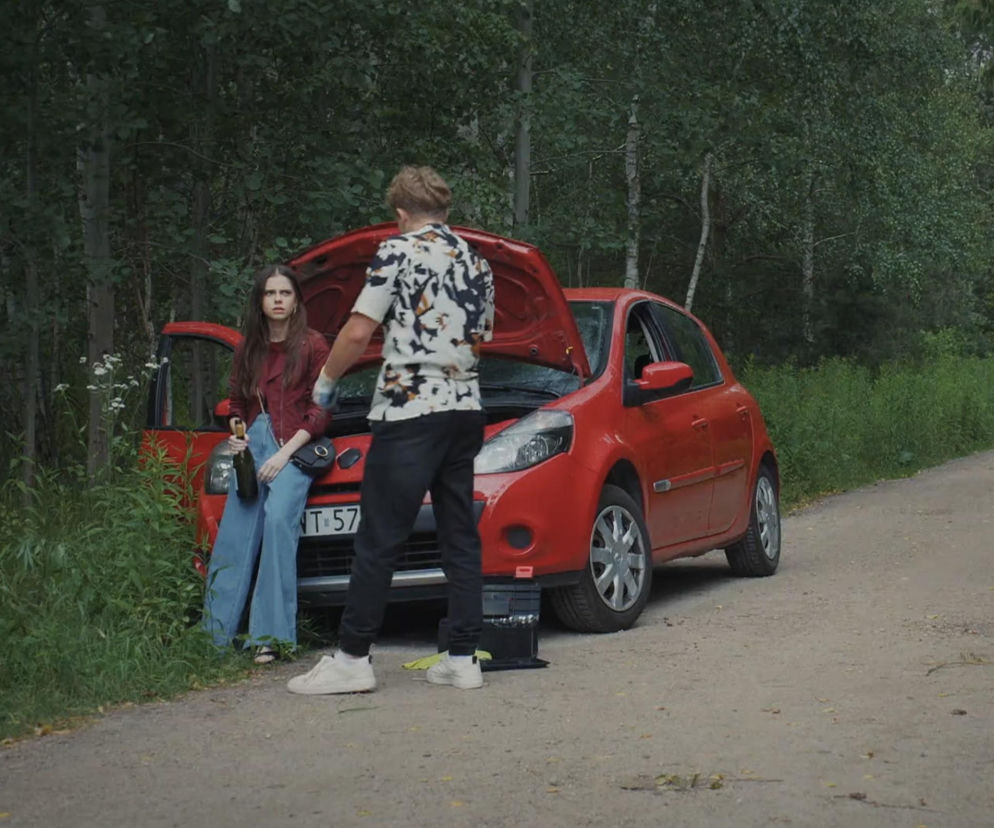 Pijana Basia (Karina Woźniak) opierająca się o zepsuty samochód z otwartą maską, trzymająca butelkę wina, obok niej Kacper (Bartosz Ziewiec) próbujący naprawić auto. Scena z serialu M jak miłość w tle lasu, o której więcej przeczytasz na Super Seriale.