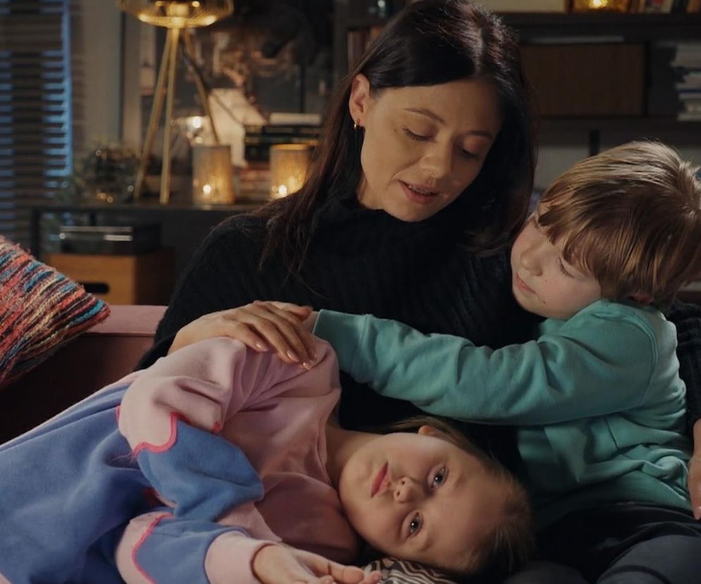 Iza Chodakowska (Adriana Kalska) z dziećmi, Majką (Laura Jankowska) i Szymkiem (Staś Szczypiński), siedzą razem na kanapie w domu. Iza spogląda w dół, trzymając rękę na Szymku, który opiera głowę na jej ramieniu. Majka leży na poduszce, patrząc prosto na widza. Więcej o dramatycznych losach Izy na Śląsku na Super Seriale.