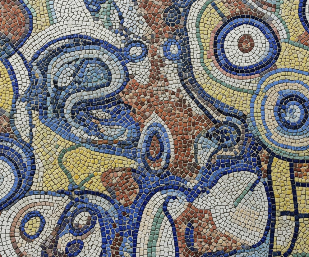 Mozaika składa się z drobnych, nieregularnych kawałków kamieni lub szkła w różnorodnych barwach, tworzących abstrakcyjny wzór. Dominują odcienie niebieskiego, żółtego, brązowego oraz białego, rozmieszczone w spirale, koncentryczne okręgi i falujące linie, które przeplatają się ze sobą. Cała powierzchnia pokryta jest tym kolorowym deseniem, a drobne elementy tworzą wrażenie tekstury i głębi.
