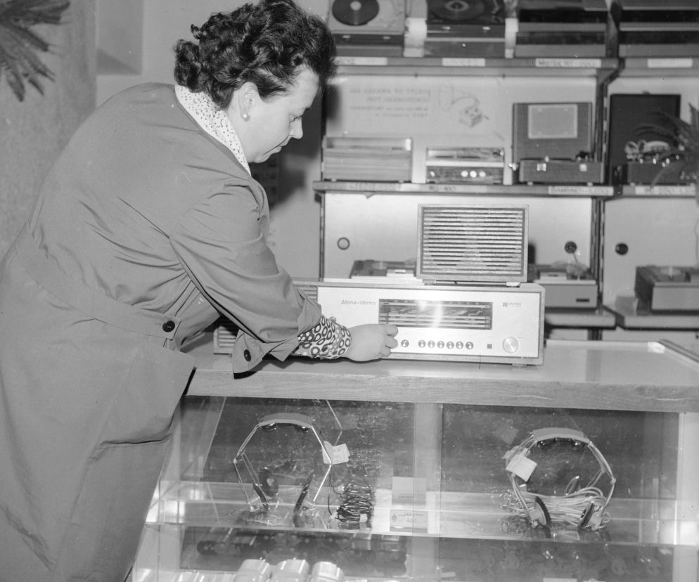 Kobieta w płaszczu dostosowuje stację radiową na sprzęcie Unitra Alena-Stereo w sklepie z elektroniką z epoki PRL. Na półkach widoczne gramofony i magnetofony, a za szybą słuchawki, to „cuda techniki” z tamtych czasów, o których przeczytasz więcej na Super Biznes.