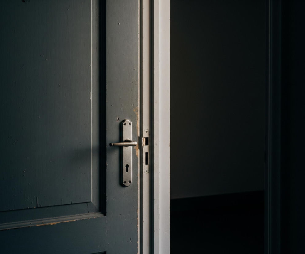 Uchylone drzwi w kolorze ciemnoszarym lub antracytowym, z lekko zdrapaną farbą na krawędziach, stanowią centrum kadru. Na ich powierzchni widać pionowe i poziome panele oraz metalową klamkę i szyld z otworem na klucz. Po prawej stronie drzwi, za uchylonym skrzydłem, widoczna jest ciemna przestrzeń, która jest praktycznie czarna, z niewielkim rozjaśnieniem u góry, sugerującym światło z innego pomieszczenia. Lewa strona obrazu jest zdominowana przez powierzchnię drzwi, która staje się ciemniejsza w kierunku lewej krawędzi.