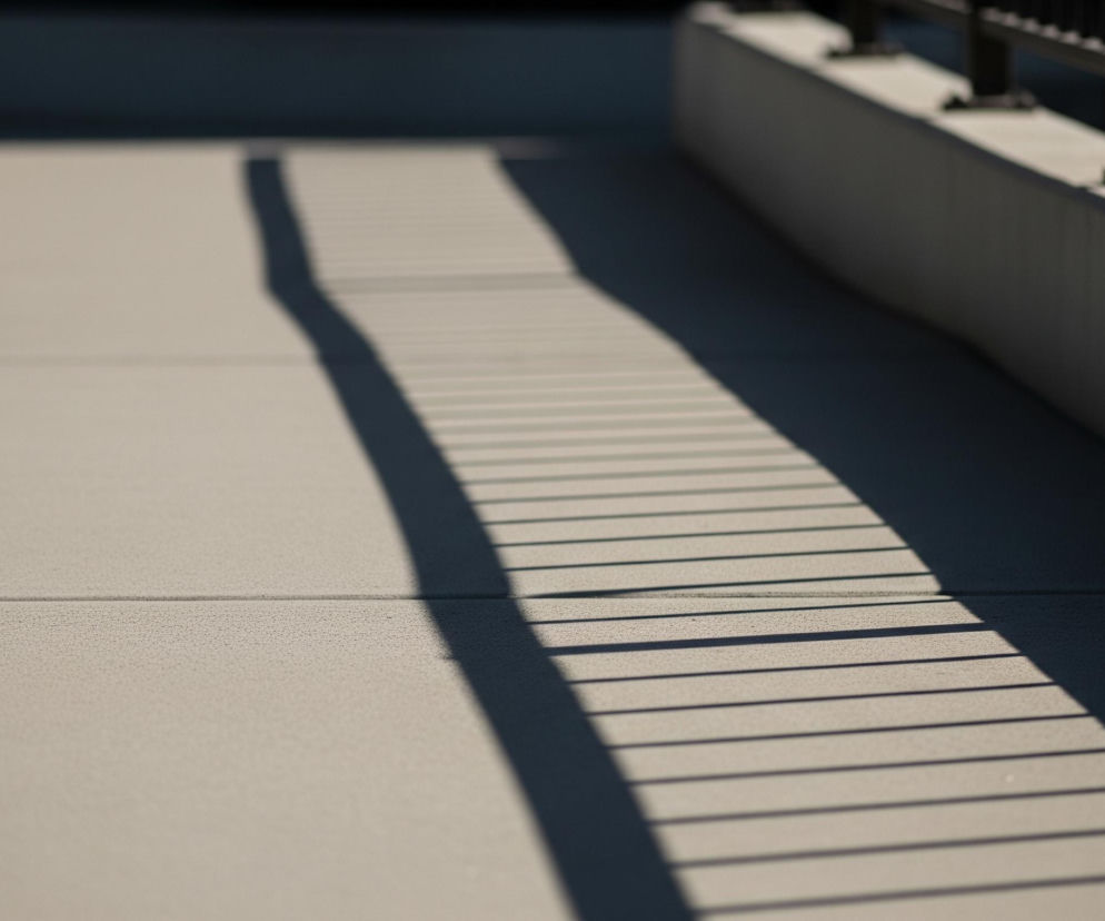 Beżowy chodnik lub podłoga z lekką teksturą zajmuje większość kadru, przecinają go dwie równoległe, ciemniejsze linie spoin. Na prawo, w górnej części obrazu, widoczna jest ciemna, metalowa balustrada z pionowymi szczeblami i poziomymi elementami konstrukcyjnymi. Od balustrady, w poprzek chodnika, rozciągają się cienie – jeden szeroki, ciemny pas i szereg równoległych, węższych cieni rzucanych przez pionowe elementy. Po lewej stronie górnego rogu widać fragment jasnej, prostej ściany.