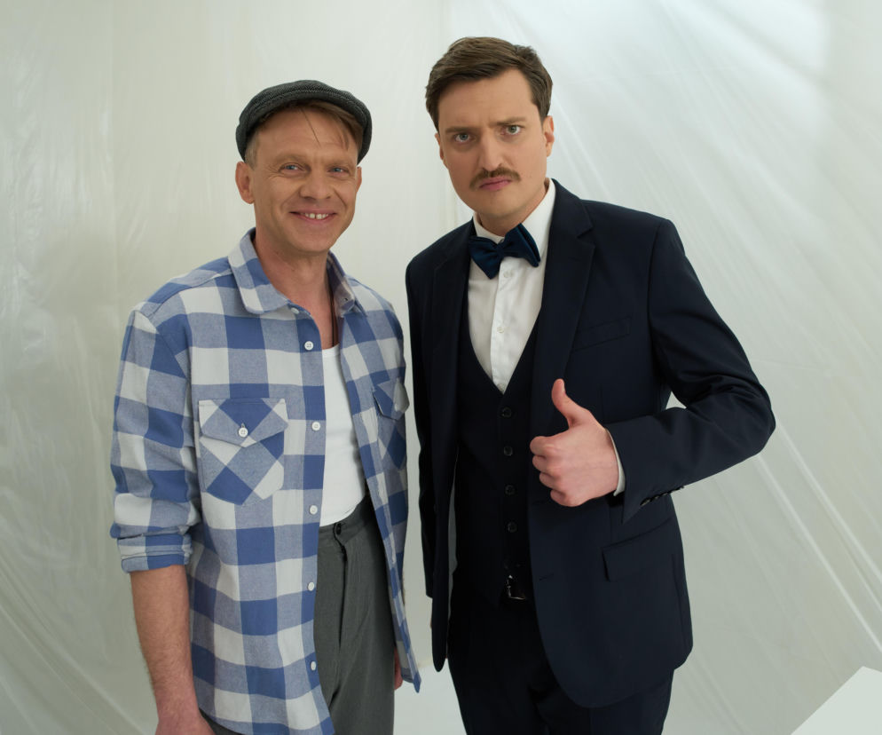 Aktorzy Cezary Sikora i Adam Woronowicz, Asystent i Taskmaster z programu „Taskmaster”, uśmiechają się do obiektywu; Sikora w czapce i kraciastej koszuli, Woronowicz w eleganckim garniturze z muszką, pokazuje kciuk w górę. Więcej informacji o programie przeczytasz na naszym portalu.