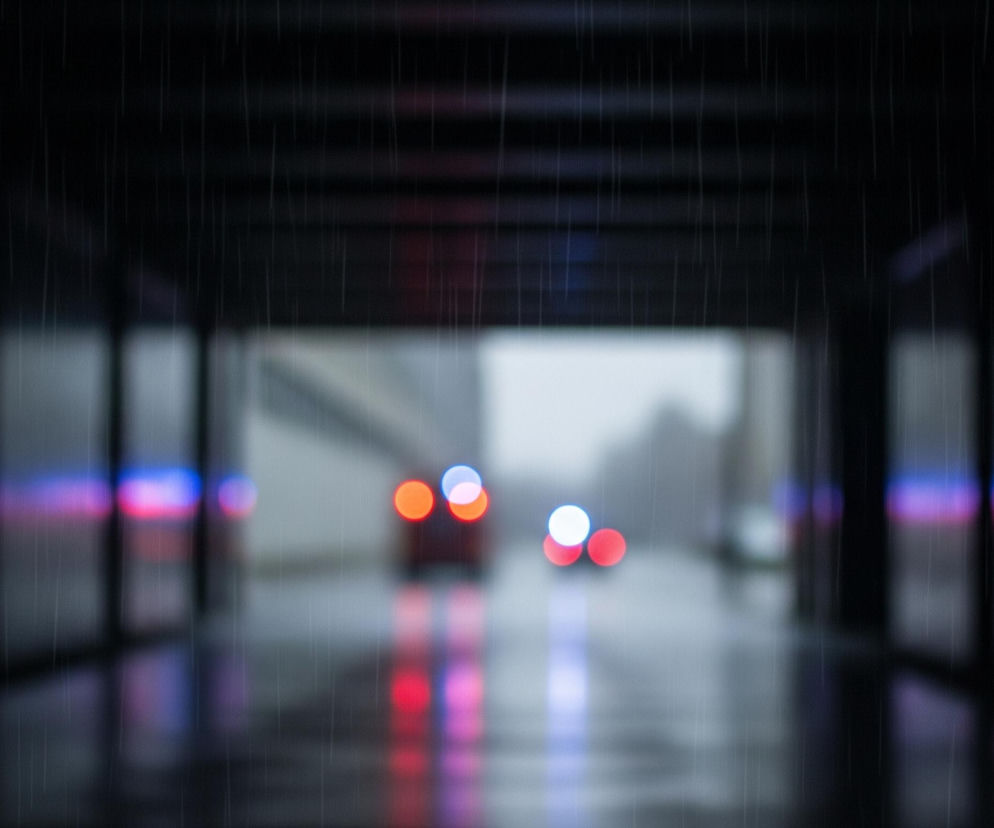 Ciemny tunel lub przejście, przez które pada deszcz, widoczny jako jasne, pionowe smugi na pierwszym planie i po bokach. W głębi, w oddali, znajdują się niewyraźne światła pojazdów, w tym dwa czerwone i dwa białe po prawej stronie oraz niebieskie i czerwone światła rozmyte po lewej stronie. Odbicia tych świateł widać na mokrej nawierzchni, tworząc podłużne, rozmyte plamy kolorów. Ciemne ściany tunelu, z widocznymi poziomymi i pionowymi elementami konstrukcyjnymi, otaczają centralne, jaśniejsze wyjście z przejścia, gdzie kolory są bardziej stonowane i szare, co sugeruje dzienną porę za tunelem.
