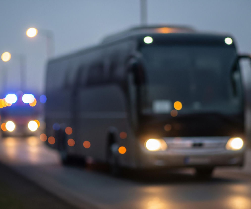 Ciemny, czarny autokar z włączonymi, jasnymi światłami przednimi i żółtymi światłami obrysowymi jedzie w dół drogi w kierunku widza, pochylony nieco w prawo. W oddali, za autokarem, widać zamazane światła innego pojazdu, prawdopodobnie niebieskie i pomarańczowe światła alarmowe. Po lewej stronie autokaru znajdują się rozmyte jasne żółte światła uliczne, a w tle widać ciemne, rozmazane sylwetki drzew lub budynków. Cała scena jest rozmyta, z wyjątkiem przedniej części autokaru, co sugeruje ruch lub płytką głębię ostrości.