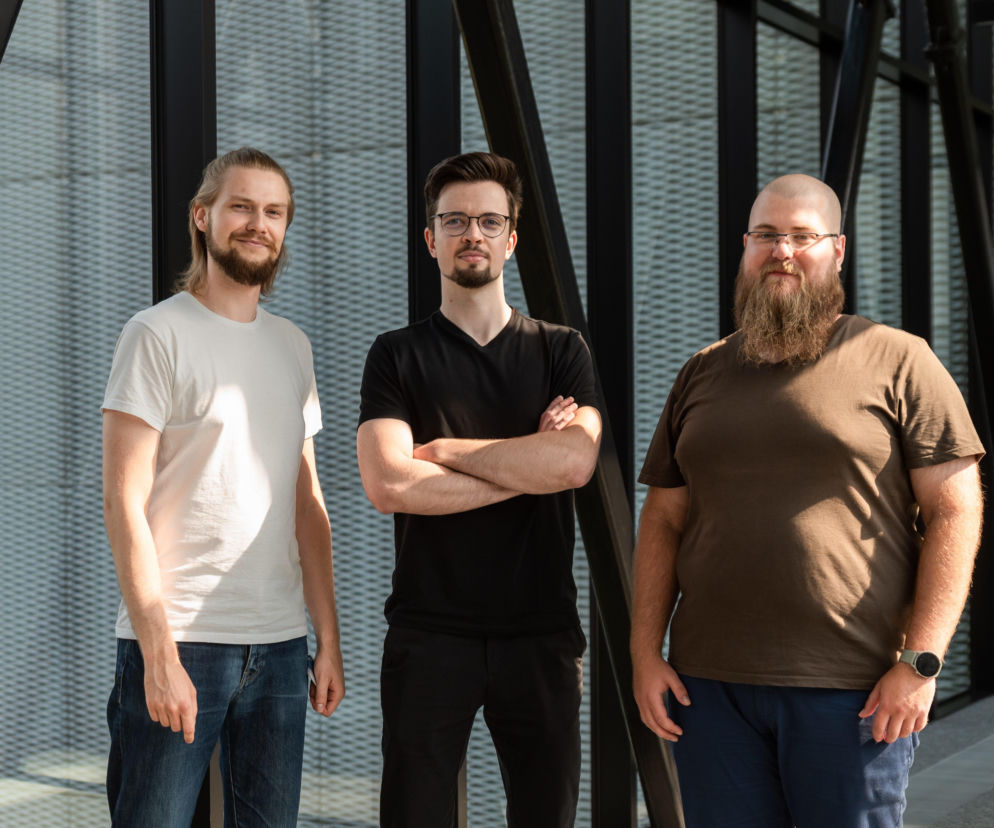 Trzech młodych mężczyzn, prawdopodobnie wynalazców lub projektantów, stojących przed nowoczesną fasadą budynku z pionowymi elementami. Uśmiechają się, prezentując pewność siebie i profesjonalizm, co wpisuje się w kontekst konkursu Jamesa Dysona, o którym przeczytasz więcej na Architektura Murator Plus.