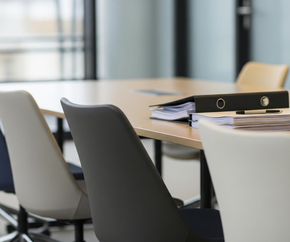 Na pierwszym planie, z prawej strony, znajduje się krzesło w jasnym odcieniu bieli, częściowo widoczne. Obok niego, nieco z lewej, stoi krzesło o ciemnoszarym obiciu, a za nim krzesło w niebieskim kolorze. Dalej w tle widać krzesło w odcieniu beżu, a za nim kolejne krzesło w kolorze niebieskim. Wszystkie krzesła ustawione są wokół długiego, drewnianego stołu o jasnej barwie. Na stole, w centralnej części, leżą stosy dokumentów i czarny segregator, a na nich widoczny jest długopis. W tle rozciąga się duże okno z czarnymi ramami, wpuszczające dzienne światło.