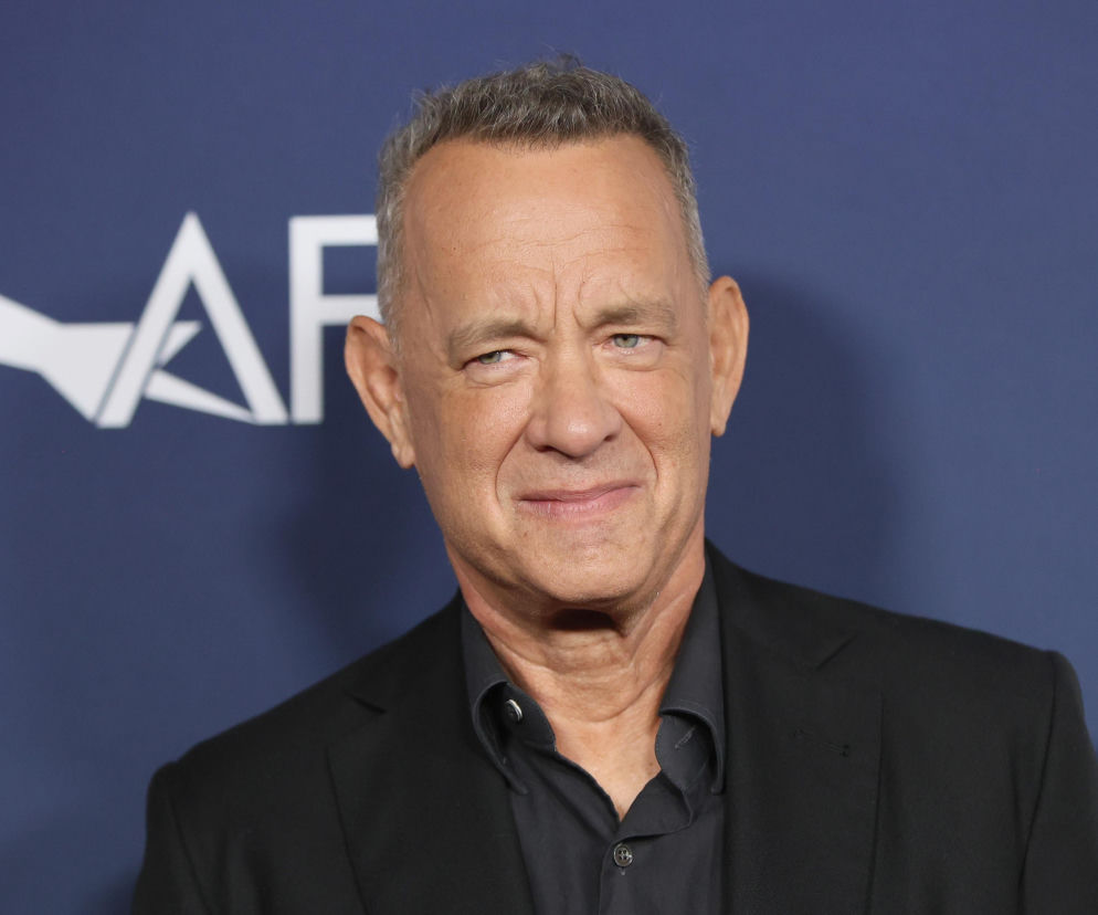 Aktor Tom Hanks w ciemnej marynarce i koszuli uśmiecha się na tle logotypu AFI. O jego pasjach przeczytasz na Eska.