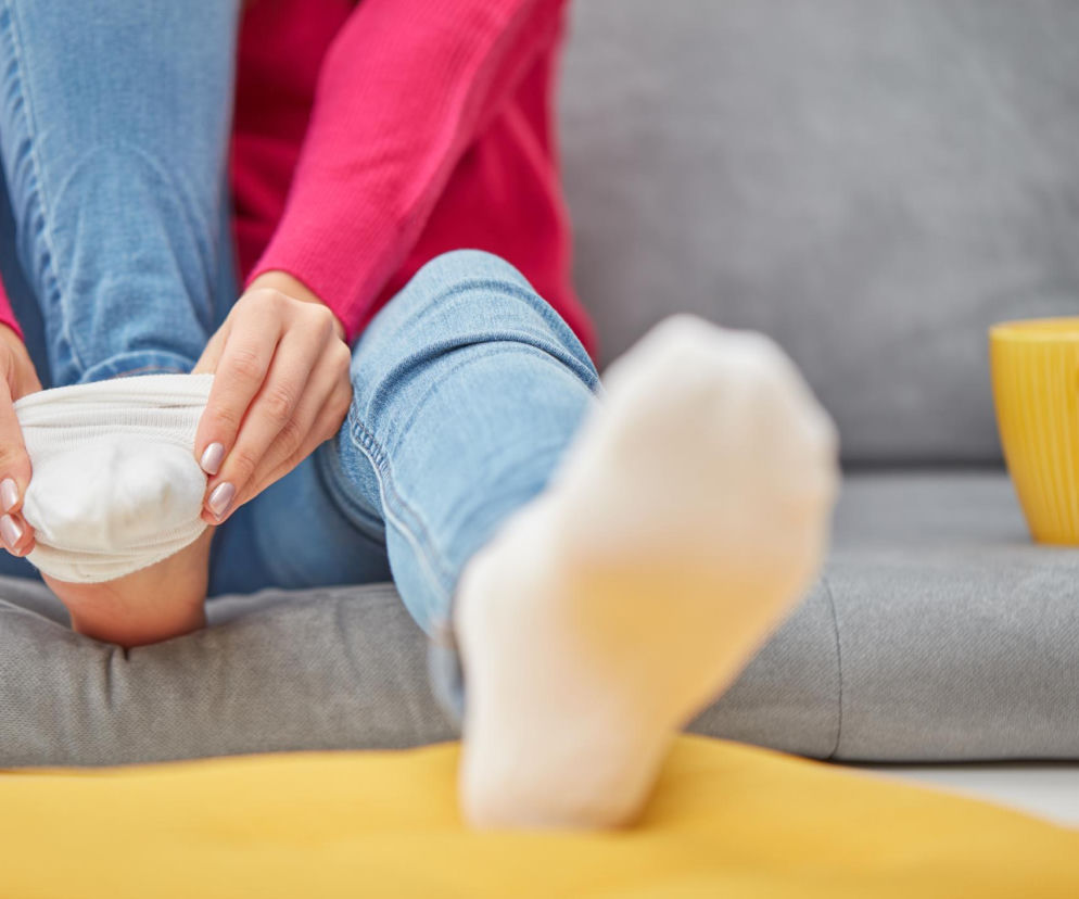 Kobieta w różowym swetrze i jeansach, siedząc na szarej kanapie, zakłada białą skarpetkę na stopę, co może być wyzwaniem przy problemach z kręgosłupem. Obok niej stoi żółty kubek. O kłopotach z mobilnością i sposobach na poprawę elastyczności ciała przeczytasz na Poradnik Zdrowie.