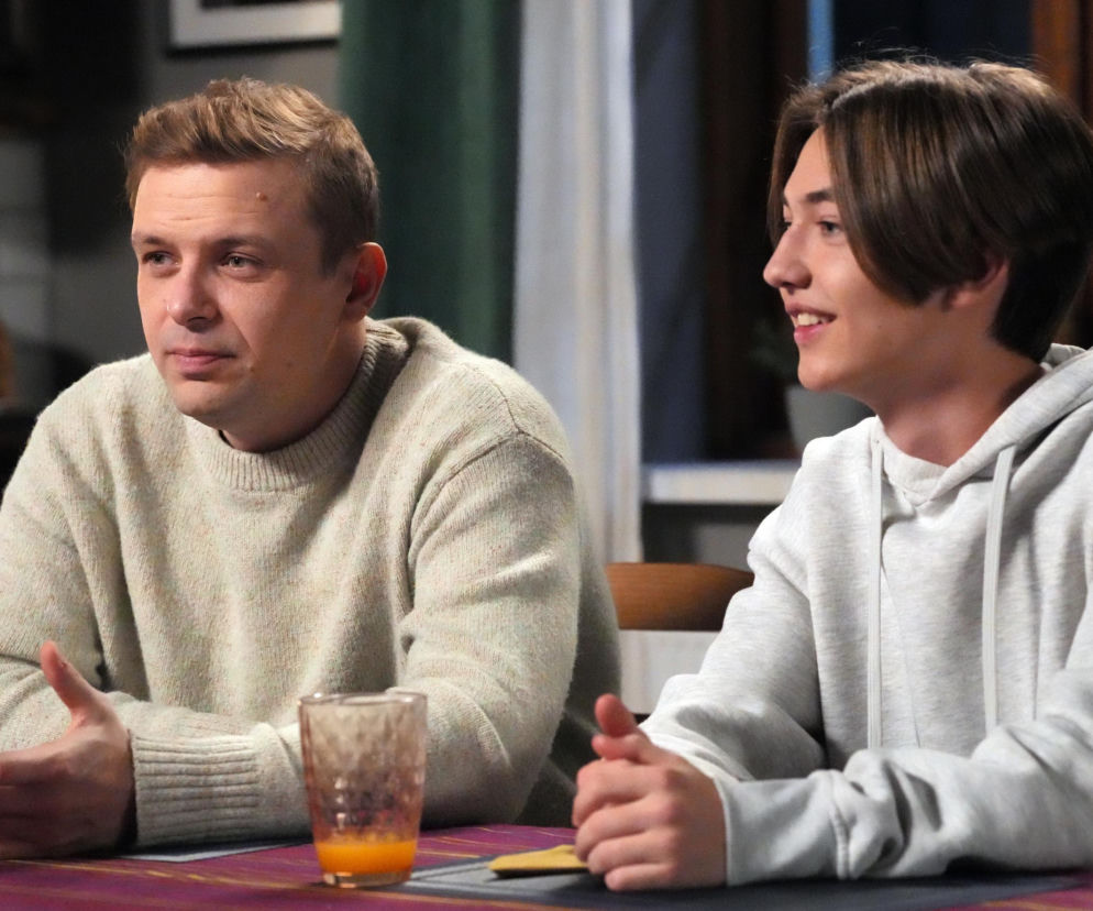 Aktorzy Marek Molak jako Hubert i Artem Malaszczuk jako Emil w serialu „Barwy szczęścia”, podczas poważnej rozmowy przy stole. Hubert, w beżowym swetrze, gestykuluje dłońmi, a Emil, w szarej bluzie z kapturem, uśmiecha się, słuchając. Więcej o ich losach na stronie Super Seriale.