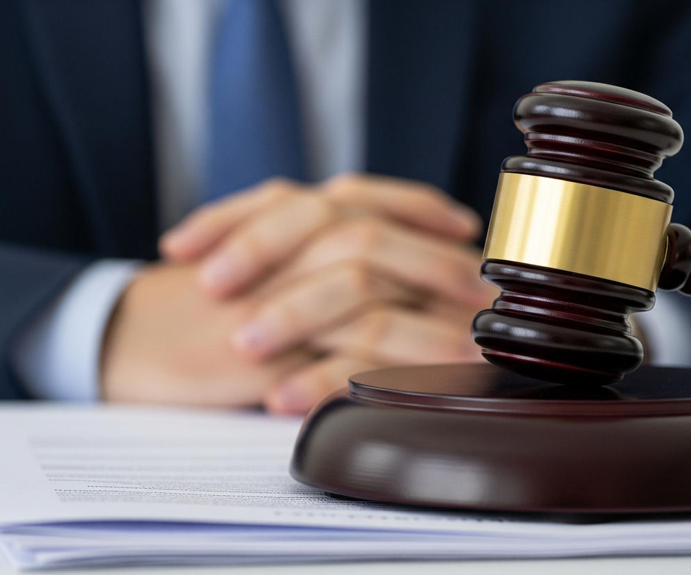 Na pierwszym planie, z prawej strony, leży ciemnobrązowy młotek sędziowski ze złotym paskiem na główce, umieszczony na pasującej, okrągłej podstawce. Pod młotkiem znajduje się stos białych kartek papieru z widocznymi drukowanymi czarnymi literami, leżący na jasnym blacie stołu. W tle, rozmyta postać osoby w ciemnoniebieskim garniturze i jasnoniebieskim krawacie, z dłońmi złożonymi na wysokości pasa, stanowi tło dla obiektów na pierwszym planie.