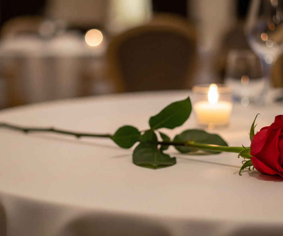 Czerwona róża leży na białym obrusie, jej długi, zielony pęd rozciąga się w lewo. Po prawej stronie róży, blisko centrum, stoi zapalona świeca w szklanym naczyniu, tworząc ciepłe, rozproszone światło. Za świecą widoczny jest przezroczysty kieliszek na wysokiej nóżce, a w tle rozmyte są kontury innych stołów i krzeseł, z kilkoma jasnymi punktami świetlnymi.