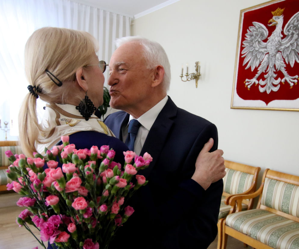 Leszek Miller i jego żona Aleksandra obejmują się serdecznie, Leszek Miller uśmiecha się i składa buziaka żonie w policzek, trzymając w tle bukiet różowych kwiatów. Biało-czerwony herb z orłem w koronie wisi na ścianie. Zdjęcie ilustruje wywiad z byłym premierem, który można przeczytać na portalu Super Biznes.
