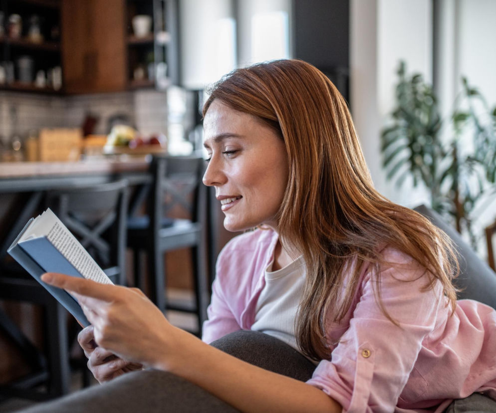 Młoda kobieta z długimi, brązowymi włosami czytająca książkę w domowym zaciszu, z delikatnym uśmiechem na twarzy. Odpoczywa na kanapie, symbolizując relaks i poznawanie tajemnic mózgu, o których przeczytasz na Poradniku Zdrowie.