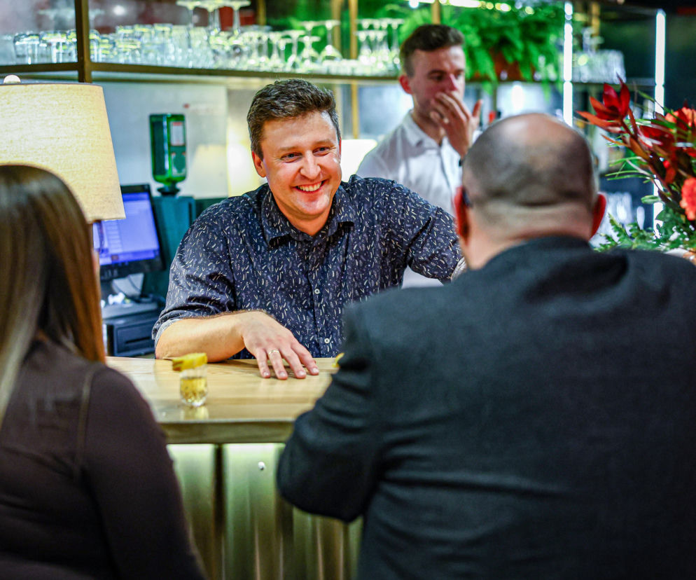 Uśmiechnięty właściciel restauracji za barem rozmawia z gośćmi. O kulisach Kuchennych Rewolucji przeczytasz na SE.
