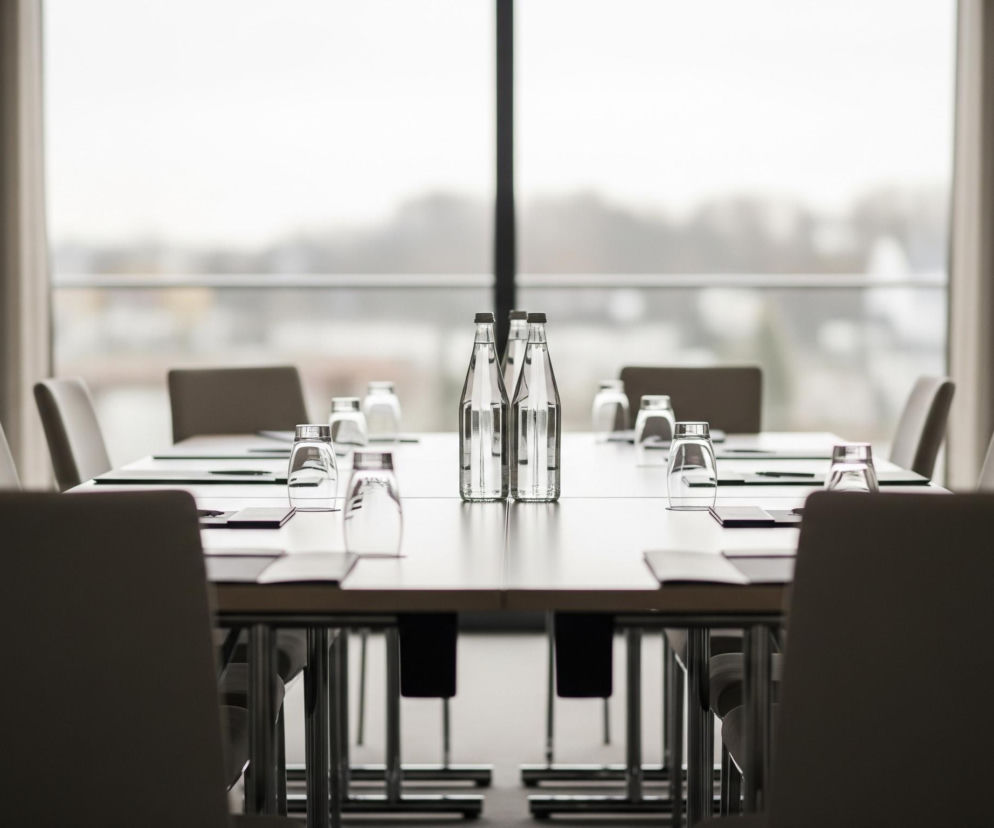 Długi, prostokątny stół konferencyjny w neutralnych odcieniach beżu i szarości zajmuje centralną część pomieszczenia. Na stole znajdują się równo ustawione szklanki oraz notatniki z długopisami, a w centralnej części stołu dwie butelki wody stoją obok siebie. Stół otoczony jest krzesłami z beżową tapicerką, widocznymi z tyłu, z których najbliższe krzesło znajduje się na pierwszym planie, częściowo zasłaniając stół. W tle rozciąga się duże okno, za którym widać rozmyty krajobraz z elementami zieleni i zabudowań, a po bokach okna wiszą beżowe zasłony.