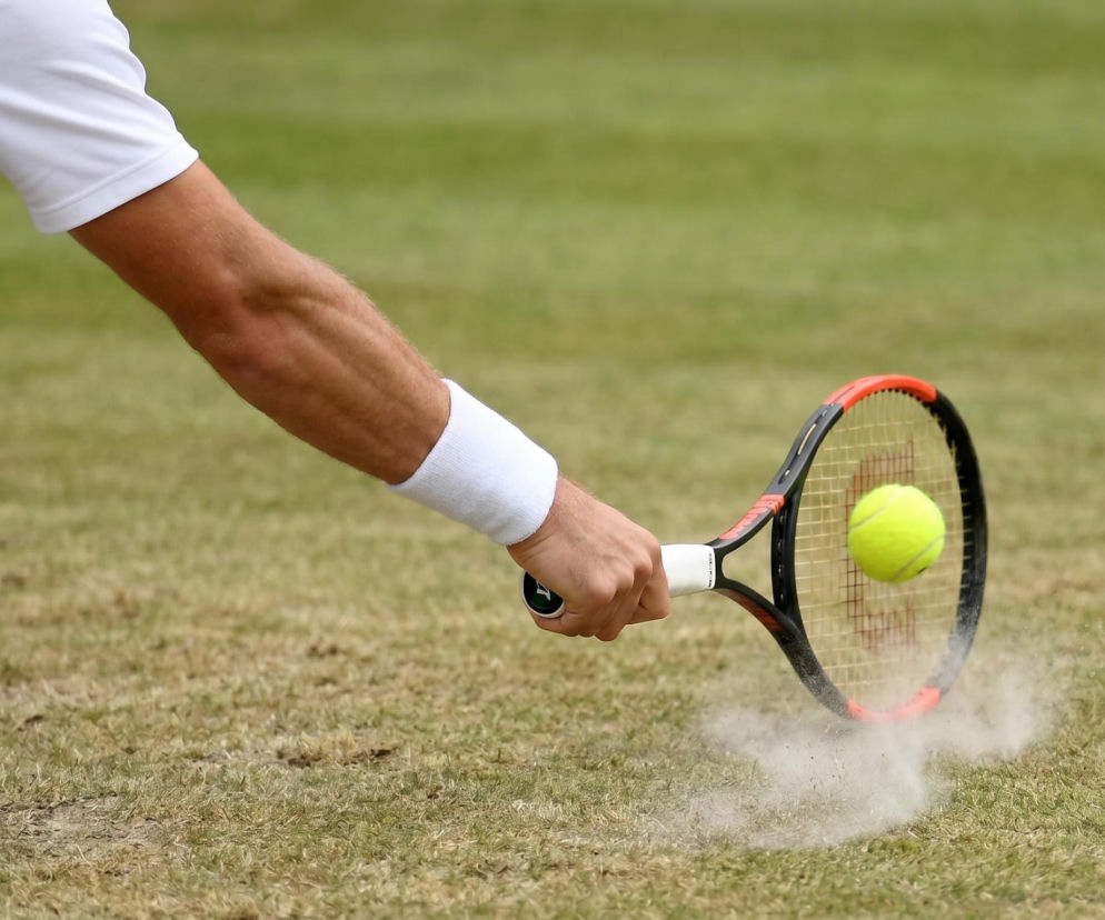 Tenisista Kamil Majchrzak w białym stroju sportowym odbija piłkę rakietą tenisową na trawiastym korcie, wzbijając chmurę pyłu. Jego mocne uderzenie symbolizuje gotowość i determinację przed Wimbledonem, o czym przeczytasz na naszym portalu.