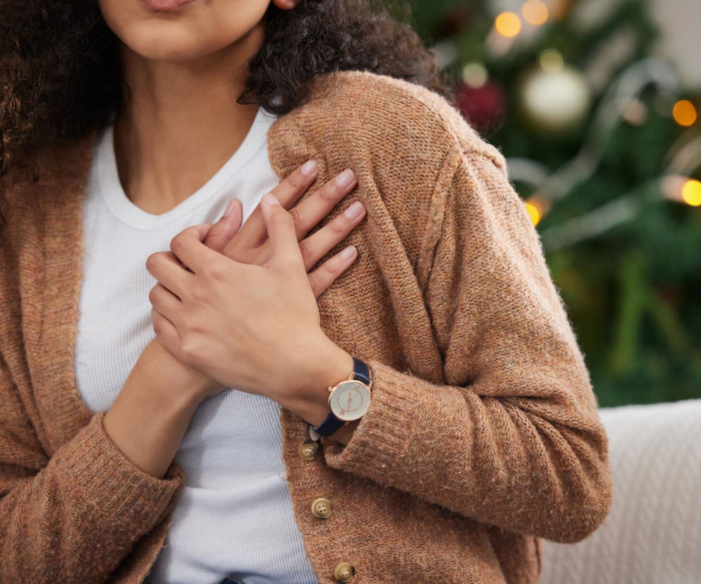 Kobieta z ciemnymi kręconymi włosami, ubrana w białą bluzkę i brązowy sweter, trzyma dłonie splecione na klatce piersiowej, sygnalizując ból w okolicy serca. W tle widać rozmazane światła choinki, co symbolizuje ryzyko zawału serca w okresie świątecznym, o czym przeczytasz na Poradnik Zdrowie.