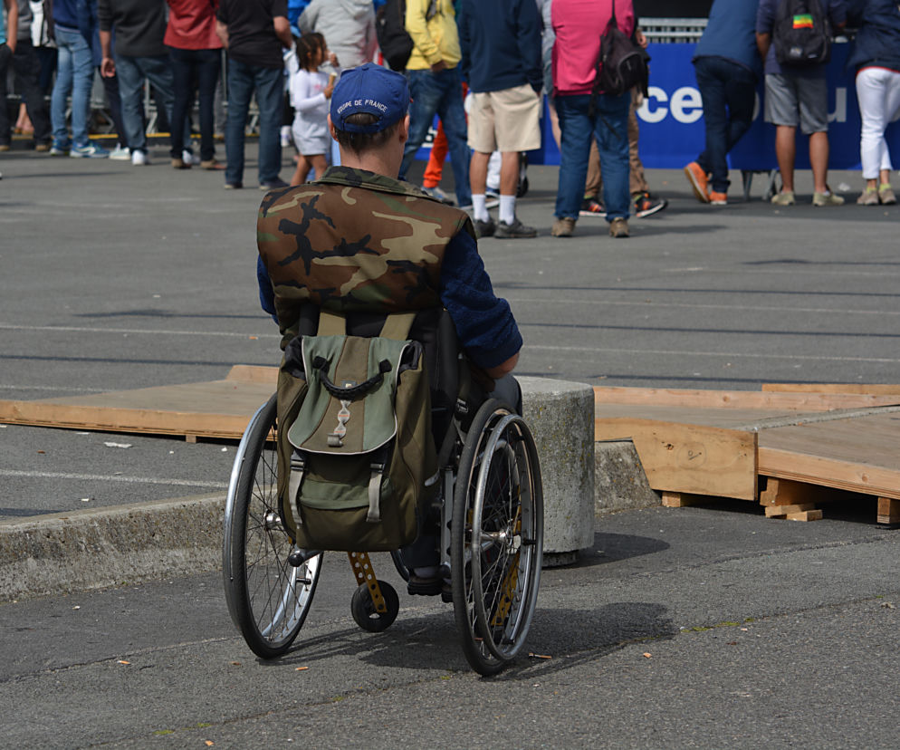 Osoba na wózku inwalidzkim z plecakiem i w czapce Equipe de France czeka na drodze obok tymczasowych drewnianych podestów. Obraz symbolizuje wyzwania i bariery, które muszą pokonywać osoby z niepełnosprawnościami. Więcej o asystencji osobistej dla niepełnosprawnych przeczytasz na Super Biznes.