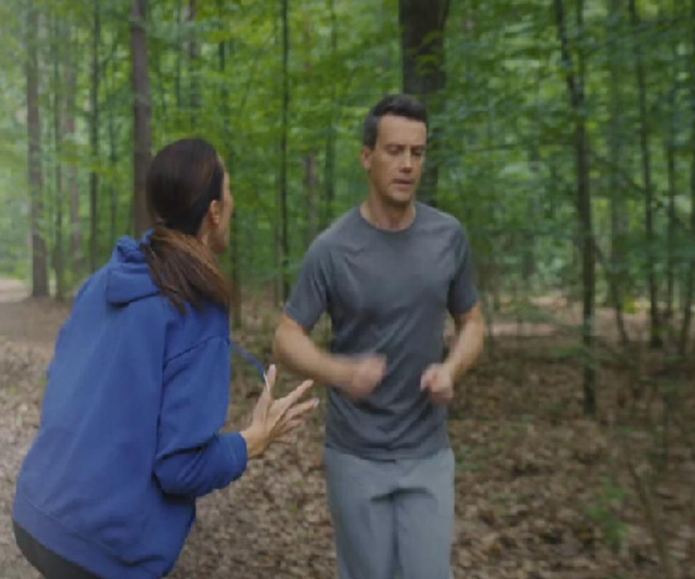 Martyna (Magdalena Turczeniewicz) w niebieskiej bluzie z kapturem, z włosami spiętymi w kucyk, gestykuluje w stronę biegnącego Jakuba (Krzysztof Kwiatkowski) w szarej koszulce i spodniach, podczas joggingu w lesie. Scena z serialu M jak miłość na Super Seriale.