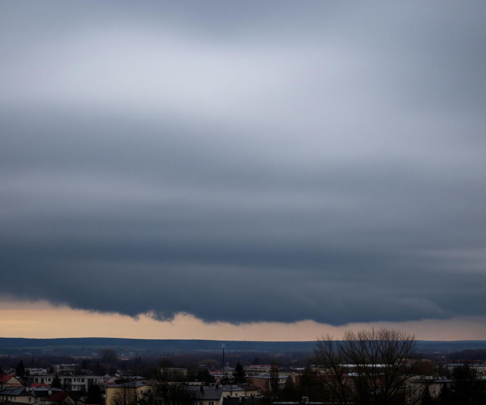 Niebo pokryte jest masywną, ciemnoszarą warstwą chmur, z jaśniejszym, niemal białym pasmem u góry, przechodzącym w ciemniejszy odcień ku dołowi. Pod chmurami rozciąga się wąski pas horyzontu w odcieniach pomarańczowo-żółtych, przechodzący w ciemny błękit krajobrazu. Na pierwszym planie widać panoramę miasta z różnobarwnymi dachami budynków, częściowo przysłoniętych nagimi drzewami i gęstym, ciemnym pasmem lasu w oddali.