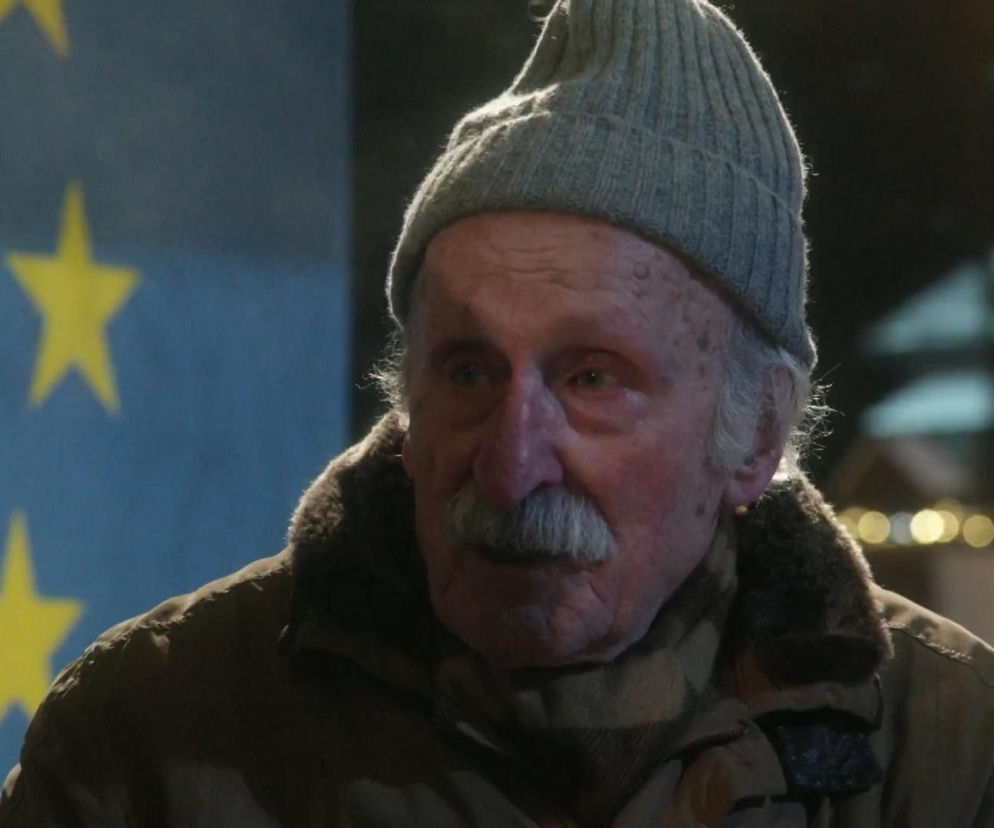 Aktor Franciszek Pieczka w roli Japycza, ubrany w szarą czapkę i kurtkę z futrzanym kołnierzem, z tłem flagi Unii Europejskiej, co symbolizuje koniec ważnej ery dla fanów serialu. Więcej o ostatnim odcinku Franciszka Pieczki w Ranczu przeczytasz na naszym portalu.
