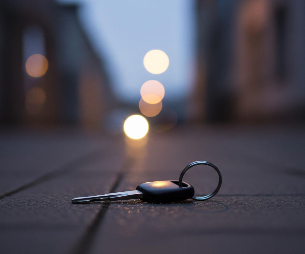 Kluczyk samochodowy z czarną obudową i metalowym ostrzem leży na ciemnej, chropowatej nawierzchni, prawdopodobnie chodniku, w perspektywie centralnej, bliżej dolnej części kadru. Klucz połączony jest z okrągłym, metalowym kółkiem. Tło jest rozmyte, ukazując niebieskawe niebo lub zmierzch oraz rozproszone, kuliste punkty światła w odcieniach żółci i bieli, prawdopodobnie uliczne latarnie. Po bokach w oddali widoczne są niewyraźne, ciemne kształty budynków.