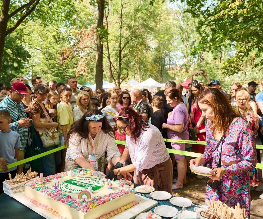 Uczestnicy Podlaskiego Śniadania Mistrzów w Ogrodach Branickich czekają na porcje tortu z logo imprezy. Dwie kobiety kroją duży, biały tort ozdobiony kolorowym kremem i małymi figurkami, podczas gdy tłum ludzi obserwuje to wydarzenie. Więcej o Podlaskim Śniadaniu Mistrzów przeczytasz na naszym portalu.