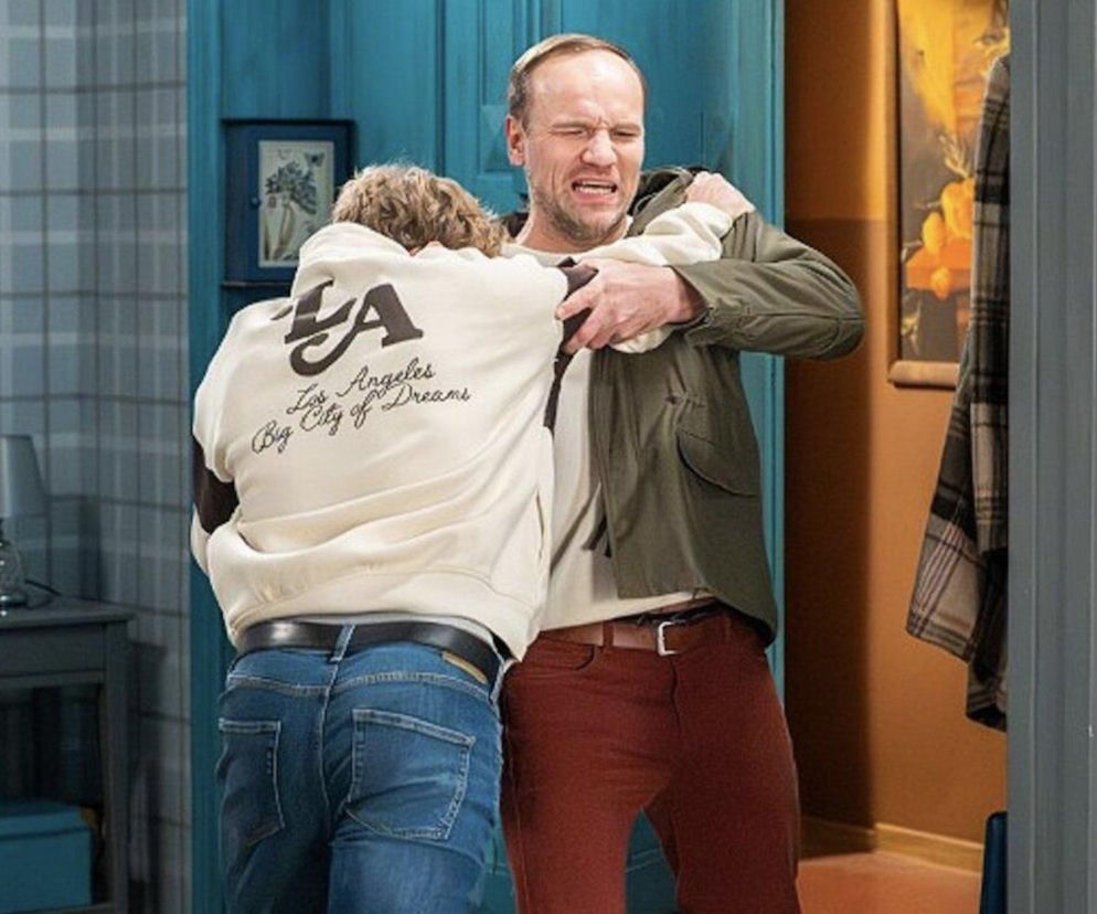 Dwóch mężczyzn, Janek i Tomek, szarpie się w mieszkaniu, tworząc dynamiczną scenę bójki. Janek w zielonej kurtce ma wykrzywioną bólem twarz, próbując powstrzymać Tomka, ubranego w beżową bluzę i dżinsy, który odwrócony jest tyłem do widza. Ten dramatyczny moment z serialu Pierwsza miłość można zobaczyć na Super Seriale.