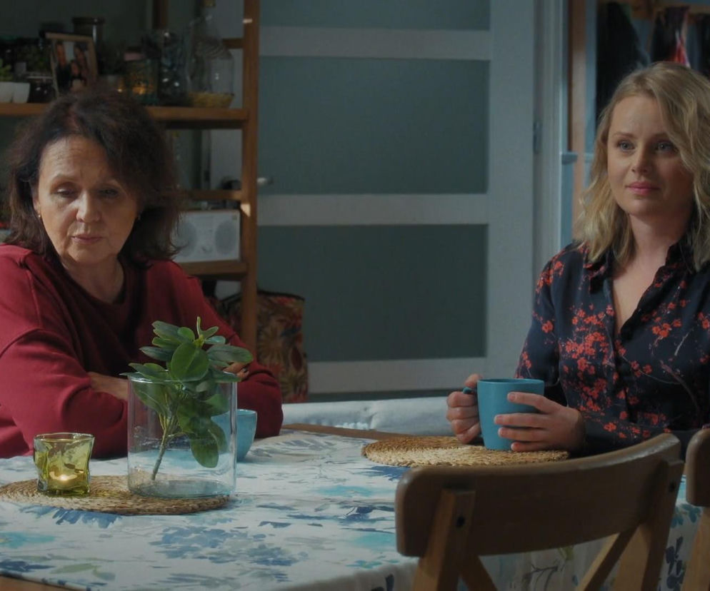 Dwie kobiety, Marysia Rogowska i Judyta Kosowska, siedzą przy stole, rozmawiając. Judyta w bluzce w kwiaty trzyma kubek, Marysia w czerwonej bluzie spogląda w dół, wyrażając zmęczenie. Obie przyjaciółki dyskutują o problemach w odcinku M jak miłość numer 1920. Więcej o tym odcinku przeczytasz na portalu Super Seriale.