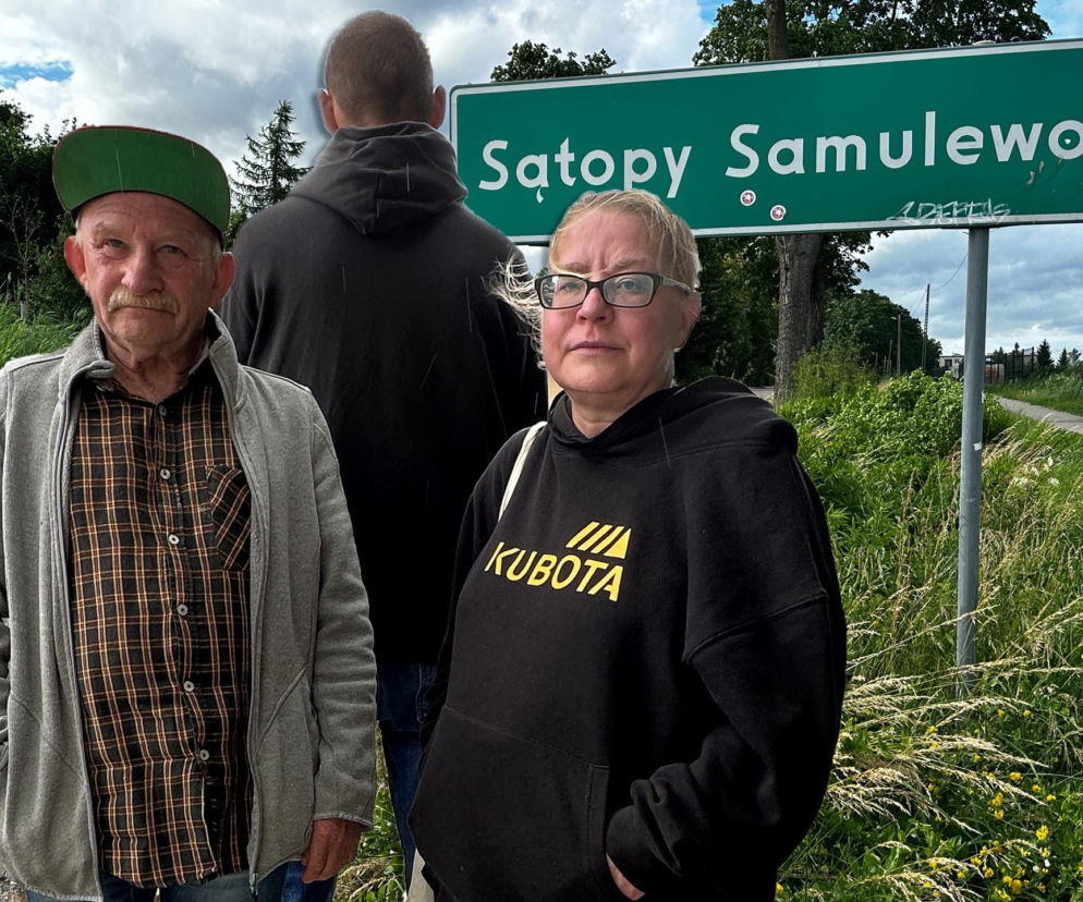 Kobieta i starszy mężczyzna na tle znaku Sątopy Samulewo. O strachu mieszkańców wsi przeczytasz na Eska Olsztyn.