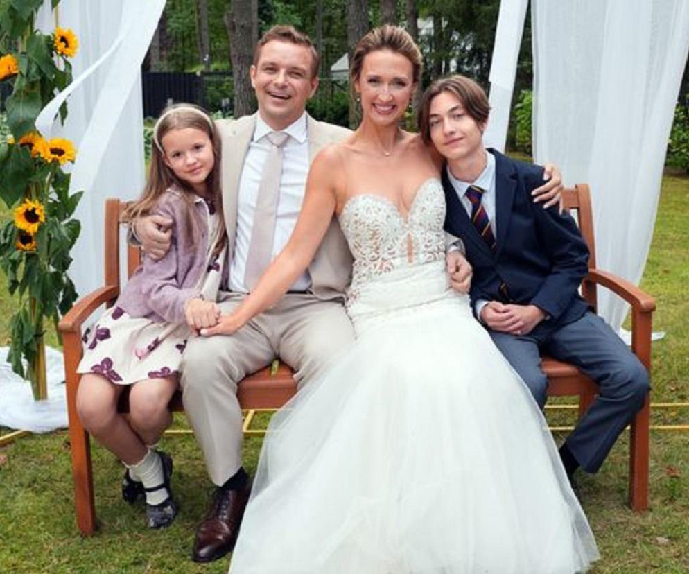 Uśmiechnięta Asia (Anna Gzyra) w białej sukni ślubnej i Hubert (Marek Molak) w beżowym garniturze z synem Emilem (Artem Małaszczuk) i Marysią (Nina Szumowska) siedzący na drewnianej ławce, w tle ślubna dekoracja z białymi wstęgami i słonecznikami. Więcej o ich ślubie znajdziesz na Super Seriale.