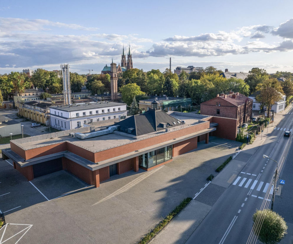 Widok z góry na Dom Pogrzebowy Koperski w Żyrardowie, ceglany budynek z latarnią doświetlającą i przeszklonym wejściem, połączony z historyczną zabudową. W tle zielone drzewa i fragment miasta, w tym kościół z wysokimi wieżami. Artykuł o tej realizacji na portalu Architektura Murator Plus.