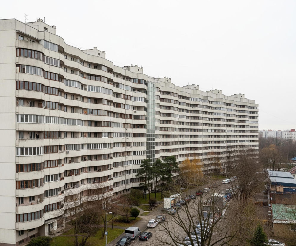 Długi, wielopiętrowy budynek mieszkalny o jasnoszarej fasadzie dominuje w centrum kadru, rozciągając się horyzontalnie przez całą jego szerokość. Budynek posiada liczne, nieregularnie rozmieszczone okna o brązowych i białych ramach oraz falujące balkony na kolejnych piętrach, nadając mu dynamiczny kształt. Przed budynkiem widoczna jest ulica z zaparkowanymi samochodami oraz skwer z drzewami o nagich gałęziach i pojedynczymi zielonymi iglakami, a dalej po prawej stronie niższe, starsze budynki z brązowymi dachami. Horyzont zamyka szereg oddalonych wieżowców o jasnych elewacjach, a nad całością rozciąga się jednolite, jasnoszare niebo.