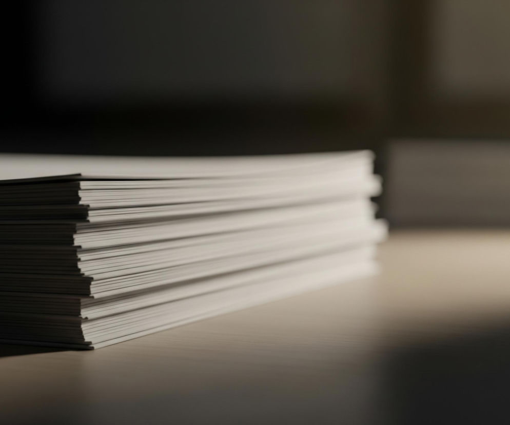 Duży stos białych kartek papieru leży na drewnianym blacie. Kartki są ułożone równo jedna na drugiej, tworząc wysoki prostopadłościan o jasnych, widocznych brzegach. Prawy górny róg stosu jest lekko zaokrąglony, co może sugerować, że są to broszury lub cienkie książki, a nie pojedyncze arkusze. Na drewnianym blacie widoczne są subtelne poziome paski. Prawa strona obrazu, za stosem papieru, jest rozmyta i pogrążona w ciemności, z ledwie widocznymi zarysami innych przedmiotów na dalszym planie. Lewa strona blatu jest również zacieniona. Światło pada z góry, oświetlając przede wszystkim górną i prawą krawędź stosu papieru oraz część blatu pod nim, tworząc łagodny cień po lewej stronie obiektu.