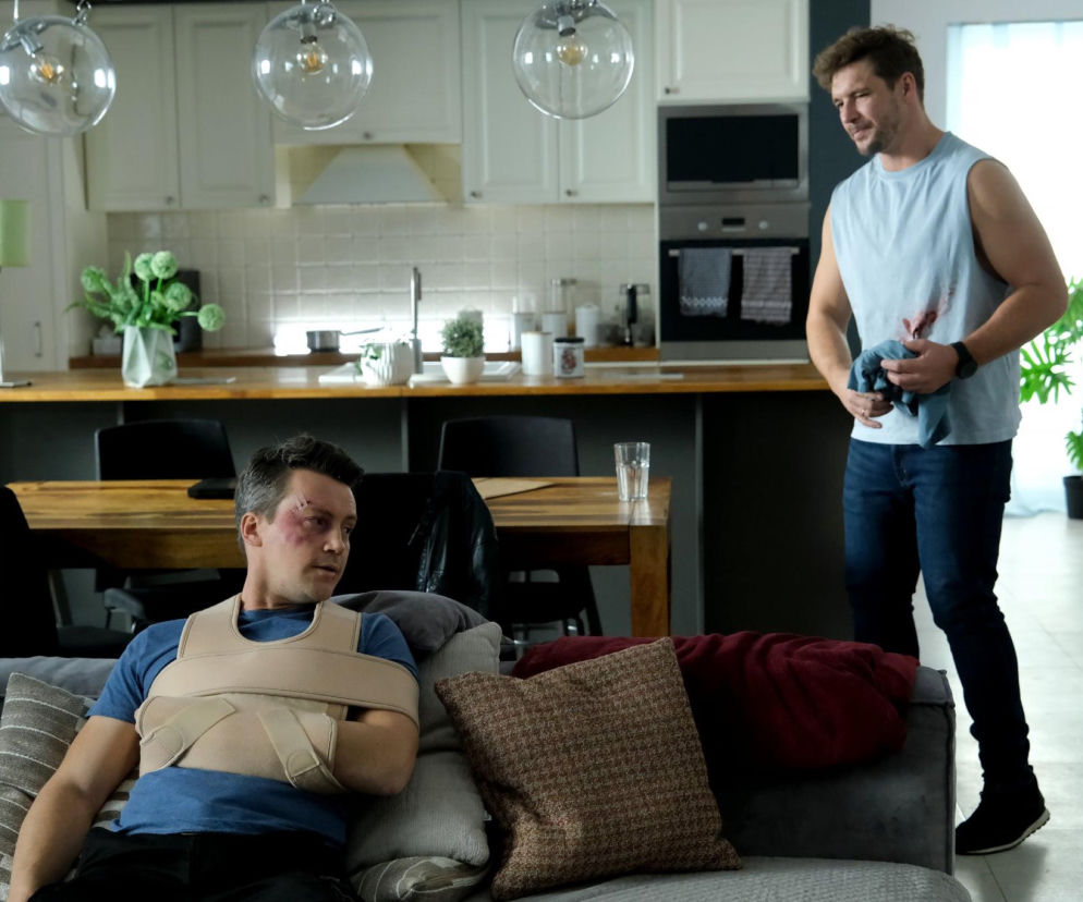 Mikołaj Roznerski jako Marcin i Krzysztof Kwiatkowski jako Jakub w scenie z serialu M jak miłość. Jakub siedzi na kanapie z temblakiem i widocznymi obrażeniami twarzy, Marcin stoi w tle z zakrwawioną koszulką. To kadr z odcinka, gdzie detektywi zostali pobici, co wywołuje kryzys w związku Marcina. Więcej na Super Seriale.