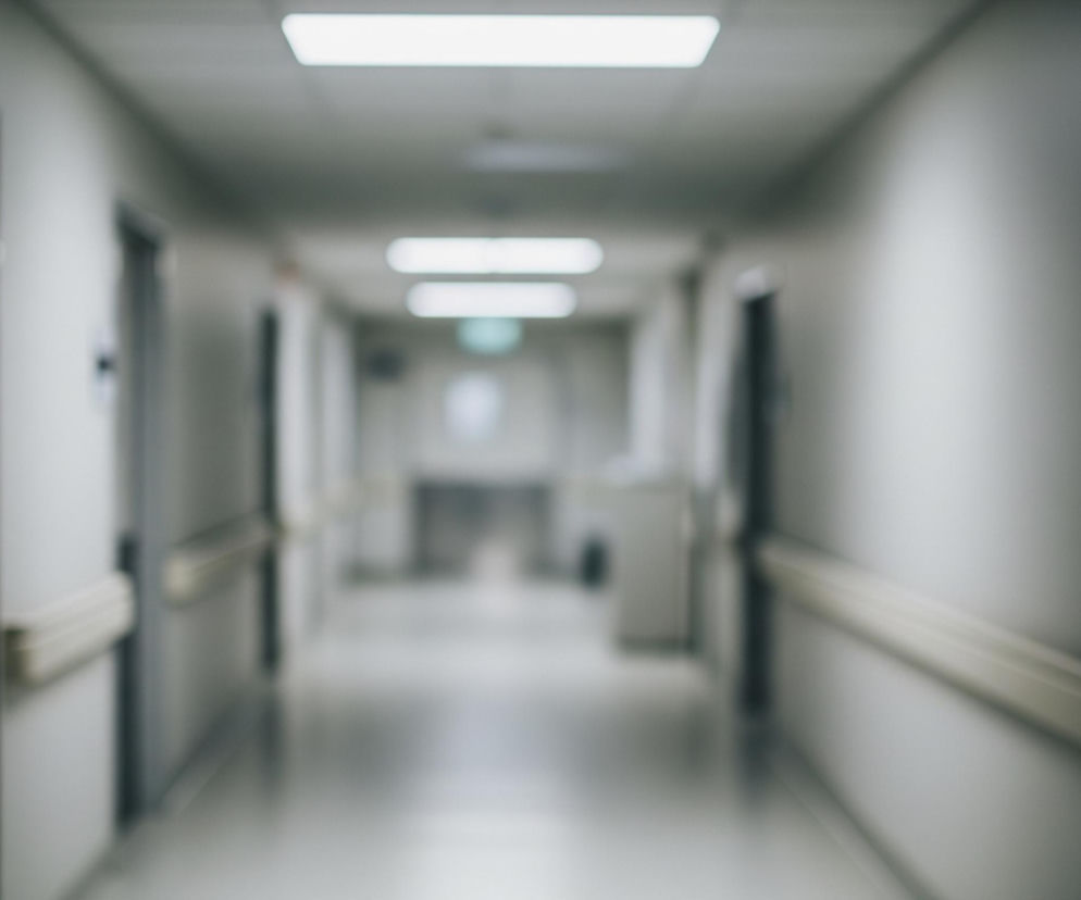 Długi, prosty korytarz rozciąga się w głąb obrazu, stopniowo zwężając się i zacierając. Po obu stronach widoczne są drzwi do pokoi, rozmieszczone w regularnych odstępach, oraz beżowe poręcze biegnące wzdłuż ścian. Sufit oświetlają prostokątne, jarzeniowe lampy, a podłoga ma jaśniejszy odcień szarości niż ściany. Cała scena jest rozmyta, co nadaje jej eteryczny i odległy charakter.