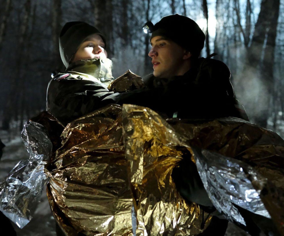 Mateusz Mostowiak, ubrany w wojskowy strój i czapkę, z latarką na głowie, niesie ranną Majkę, okrytą folią termiczną, w ciemnym lesie nocą, z dymkiem pary unoszącym się z ust. W tle niewyraźna postać z włączoną czołówką. Więcej o dramatycznym finale poszukiwań dziecka przeczytasz na Super Seriale.