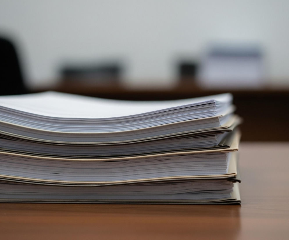 Na pierwszym planie, z lewej strony, widać ułożony stos czterech segregatorów z papierowymi dokumentami. Segregatory mają beżowe okładki i są wypełnione białymi kartkami, które delikatnie wystają poza krawędzie oprawy, tworząc faliste linie. Obiekty te leżą na brązowym, błyszczącym blacie biurka, który odbija światło. Tło jest rozmyte, ukazując niewyraźne kształty: ciemny, czarny obiekt po lewej stronie, być może oparcie krzesła, oraz jaśniejszą ścianę za nim, a także dalsze, nieostre przedmioty na biurku, takie jak biały obiekt, co sugeruje wnętrze biura.