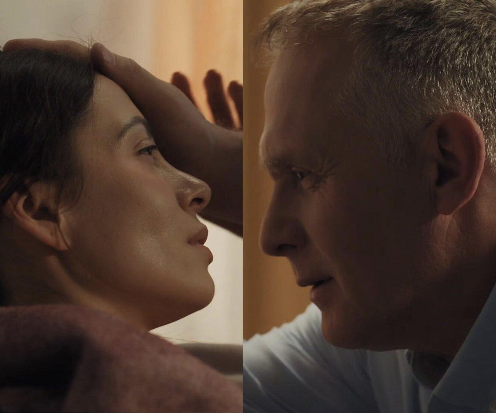 Zbliżenie na twarz Joanny (Dominika Sakowicz) w tle z dłonią Artura Rogowskiego (Robert Moskwa), który troskliwie sprawdza jej temperaturę oraz profil Artura w jasnej koszuli, patrzącego na chorą. Scena z serialu M jak miłość o początkach intymnej relacji i chorobie Joanny na Super Seriale.