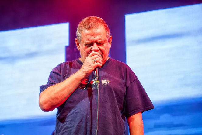Kazik Staszewski, lider zespołu Kult, śpiewa podczas koncertu, trzymając mikrofon i z zamkniętymi oczami, co sugeruje intensywność występu. Artysta, ubrany w ciemną koszulkę, mierzy się z problemami zdrowotnymi, o czym możesz przeczytać na portalu Poradnik Zdrowie.