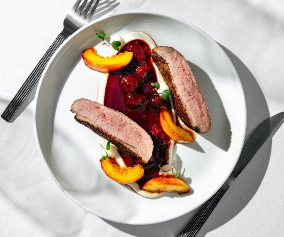 Elegancko podana pierś z kaczki z chrupiącą skórką, sosem wiśniowym, plastrami brzoskwini i kremowym purée, udekorowana świeżą zieleniną na białym talerzu, obok widelca i noża. Odkryj najlepsze restauracje na FineDiningWeek, czytając artykuł na naszym portalu.
