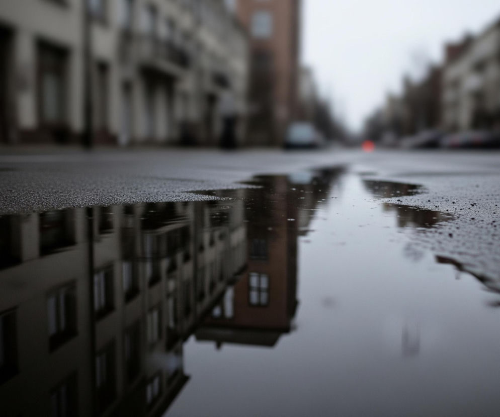 Na pierwszym planie widoczna jest duża kałuża wypełniająca część ulicy, której powierzchnia odbija fasady budynków po lewej stronie kadru. Asfalt ulicy, pokryty drobnymi, ciemnymi kamyczkami, jest mokry i błyszczący. W tle, wzdłuż obu stron jezdni, rozciągają się rozmazane fasady kamienic o stonowanych barwach szarości i beżu, z widocznymi oknami i balkonami. Na horyzoncie, również niewyraźne, rysują się kontury zaparkowanych samochodów oraz pojedyncze, niewyraźne czerwone światło.