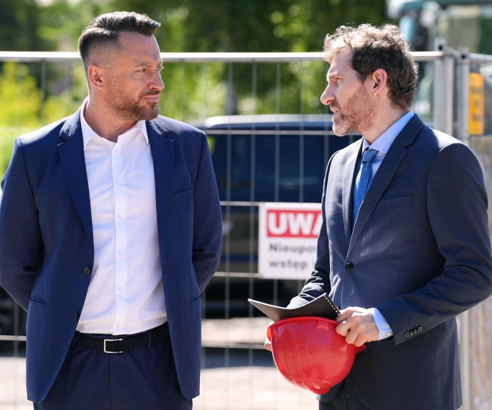 Maciej Wilk (Adam Szczyszczaj) i Szczepan (Robert Gulaczyk) w garniturach stoją na placu budowy. Wilk, z rękoma założonymi za plecami, patrzy na Szczepana trzymającego czerwony kask. Widoczny baner UWAGA Nieupoważnionym wstęp wzbroniony. Więcej o ich losach na Super Seriale.