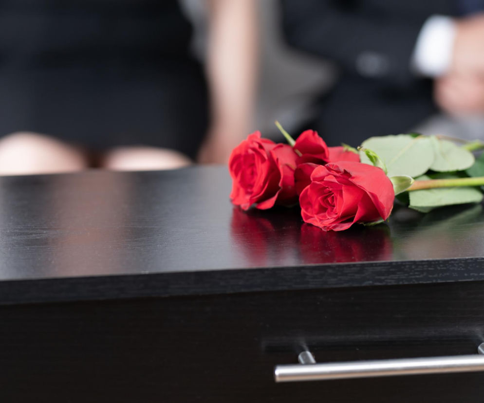 Dwie czerwone róże leżące na ciemnej, drewnianej trumnie, z rozmazanymi postaciami żałobników w tle, symbolizujące dramatyczne wydarzenie, gdzie skrajna hipoglikemia może doprowadzić do pomyłki ze zgonem, o czym można przeczytać na portalu Poradnik Zdrowie.